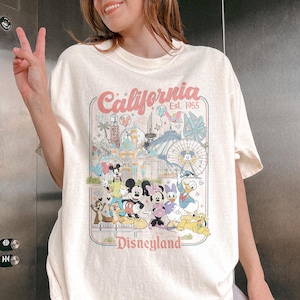 May include: Cream-colored t-shirt featuring a colorful graphic of Disneyland characters and attractions. The design includes the text "California Est. 1955" and "Disneyland" in a retro style. The shirt is a relaxed fit.