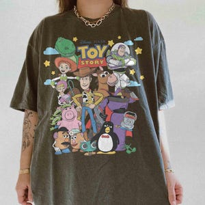 May include: Dark gray t-shirt featuring a colorful graphic of characters from the Disney Pixar movie, Toy Story. The design includes Woody, Buzz Lightyear, and other beloved characters. The shirt has a crew neck and short sleeves.