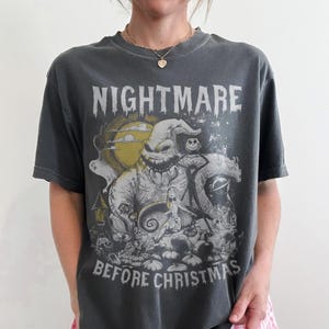 May include: Dark gray t-shirt featuring a graphic from "The Nightmare Before Christmas." The design includes the text "NIGHTMARE BEFORE CHRISTMAS" above and below a detailed illustration of characters and scenes from the movie. The shirt is worn with pink and white checkered shorts.