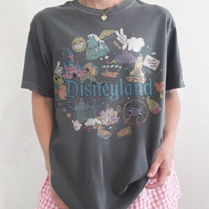 May include: Dark gray t-shirt with a colorful Disneyland graphic. The design features iconic park imagery and the word "Disneyland" in a whimsical font. The shirt is paired with pink and white checkered shorts.