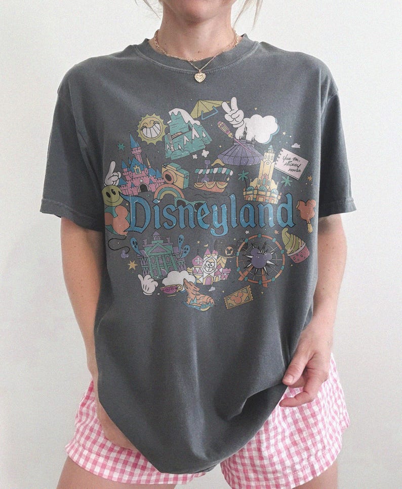 May include: Dark gray t-shirt with a colorful Disneyland graphic. The design features iconic park imagery and the word "Disneyland" in a whimsical font. The shirt is paired with pink and white checkered shorts.
