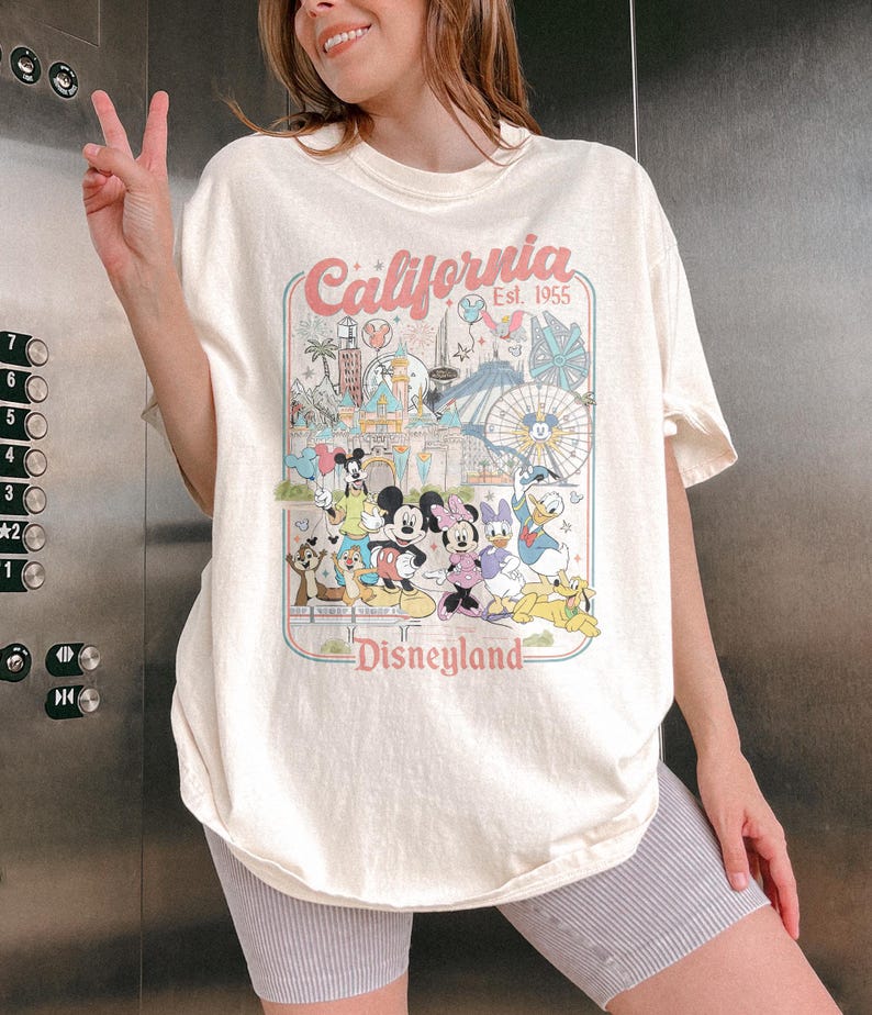 May include: Cream-colored t-shirt featuring a colorful graphic of Disneyland characters and attractions. The design includes the text "California Est. 1955" and "Disneyland" in a retro style. The shirt is a relaxed fit.