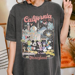 May include: Dark gray t-shirt featuring a colorful graphic of Disneyland characters and attractions. The design includes the text "California Est. 1955" and "Disneyland" in a retro style. The shirt has a vintage, worn look.