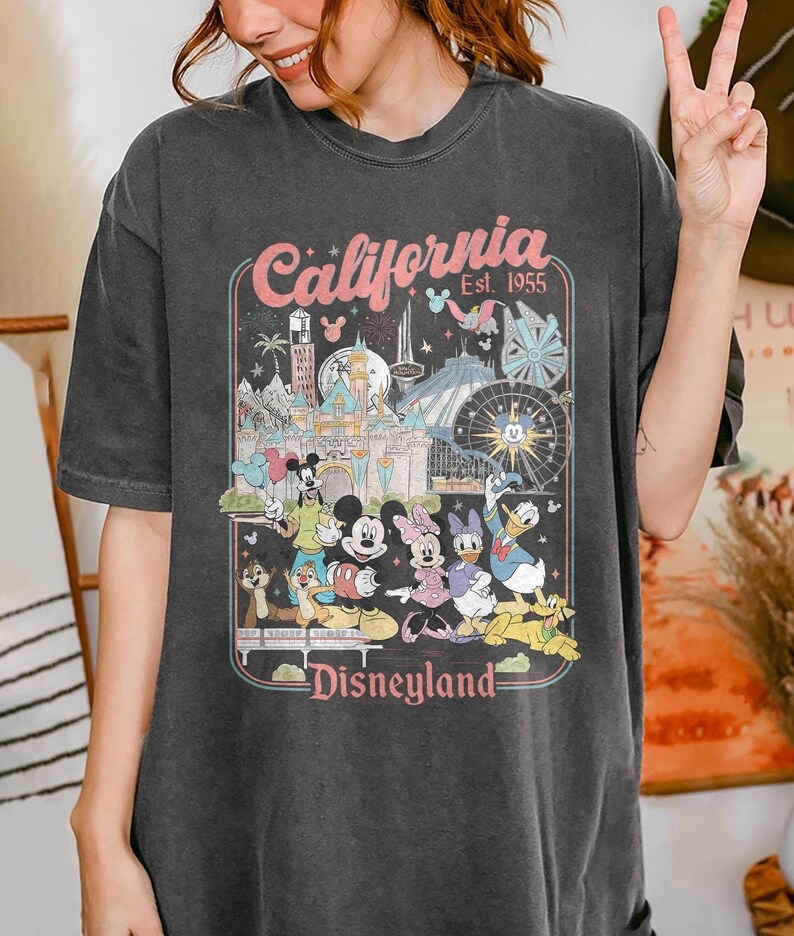 May include: Dark gray t-shirt featuring a colorful graphic of Disneyland characters and attractions. The design includes the text "California Est. 1955" and "Disneyland" in a retro style. The shirt has a vintage, worn look.