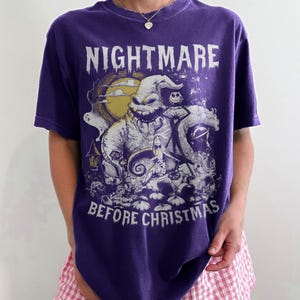 May include: A purple t-shirt featuring a graphic from "The Nightmare Before Christmas." The design includes the text "NIGHTMARE BEFORE CHRISTMAS" and illustrations of characters like Oogie Boogie and Sally. The shirt has a crew neck and short sleeves.
