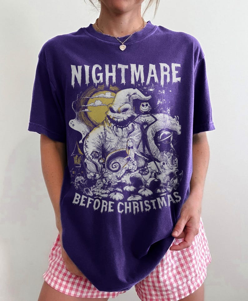 May include: A purple t-shirt featuring a graphic from "The Nightmare Before Christmas." The design includes the text "NIGHTMARE BEFORE CHRISTMAS" and illustrations of characters like Oogie Boogie and Sally. The shirt has a crew neck and short sleeves.