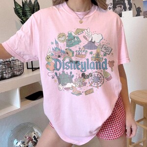 May include: A light pink t-shirt featuring a colorful Disneyland graphic. The design includes a castle, a Ferris wheel, and the word "Disneyland" in a whimsical font. The shirt is a short-sleeved crew neck.