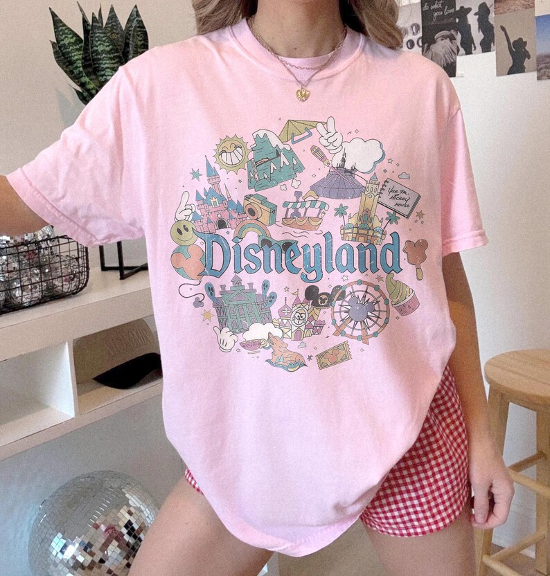 May include: A light pink t-shirt featuring a colorful Disneyland graphic. The design includes a castle, a Ferris wheel, and the word "Disneyland" in a whimsical font. The shirt is a short-sleeved crew neck.