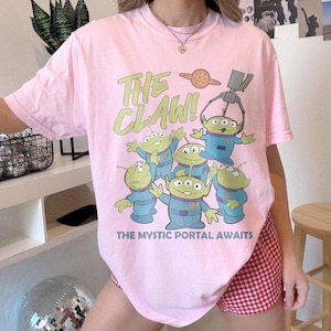 Puede incluir: Una camiseta rosa claro con un gr&aacute;fico de cinco alien&iacute;genas verdes de tres ojos con uniformes azules. Arriba, el texto dice THE CLAW! con un brazo de m&aacute;quina de garra y un planeta. Debajo, el texto dice THE MYSTIC PORTAL AWAITS.