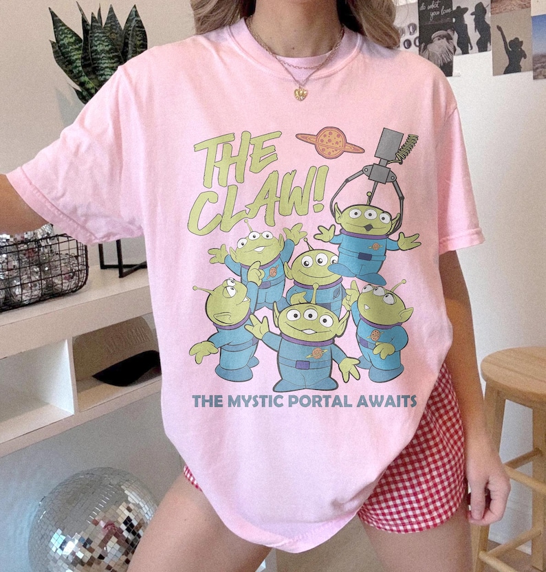 Puede incluir: Una camiseta rosa claro con un gr&aacute;fico de cinco alien&iacute;genas verdes de tres ojos con uniformes azules. Arriba, el texto dice THE CLAW! con un brazo de m&aacute;quina de garra y un planeta. Debajo, el texto dice THE MYSTIC PORTAL AWAITS.