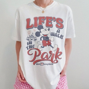 May include: White t-shirt featuring a retro design with Mickey Mouse and the text "LIFE'S A WALK IN THE PARK" in red and blue. The Walt Disney World logo is at the bottom. The shirt is a classic, casual style.
