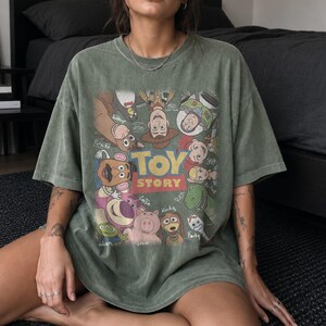 May include: An olive green short-sleeve t-shirt featuring a graphic print of Toy Story characters including Woody, Buzz Lightyear, Jessie, Rex, and others arranged around the central yellow and red Toy Story logo with character names written.