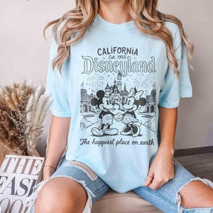 May include: Pale blue t-shirt with a black and white Disneyland graphic featuring Mickey and Minnie Mouse. The shirt displays the text "CALIFORNIA Disneyland Est. 1955" and "The happiest place on earth."