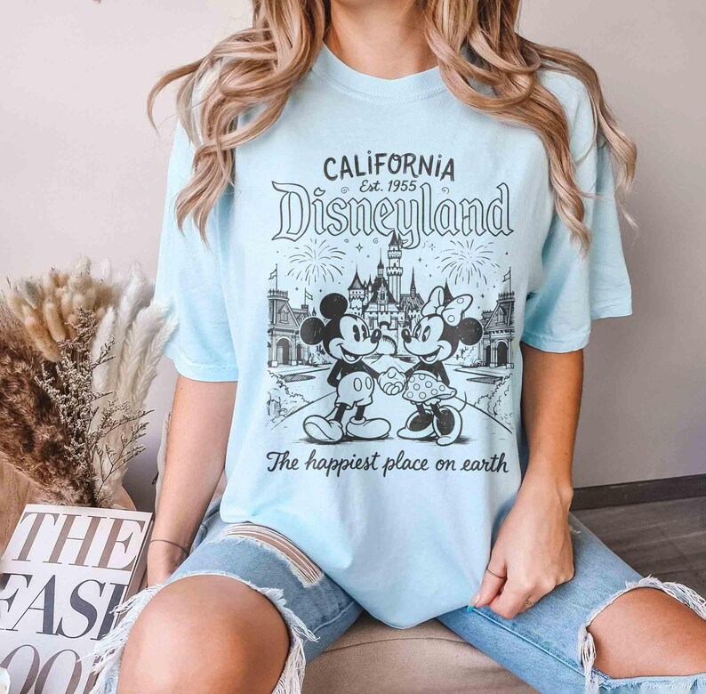 May include: Pale blue t-shirt with a black and white Disneyland graphic featuring Mickey and Minnie Mouse. The shirt displays the text "CALIFORNIA Disneyland Est. 1955" and "The happiest place on earth."