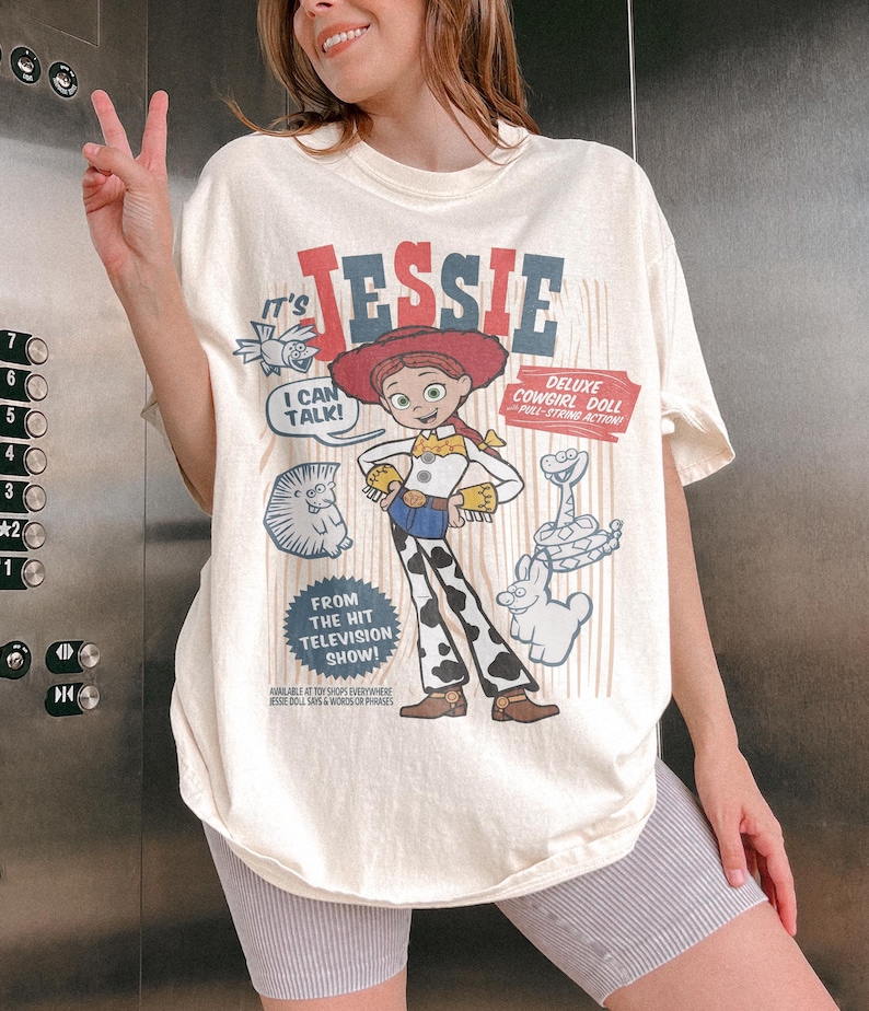 May include: Cream-colored t-shirt featuring a graphic of the character Jessie from the animated film Toy Story. The design includes the text "It's Jessie," "Deluxe Cowgirl Doll," and "From the Hit Television Show!" The shirt is paired with gray shorts.