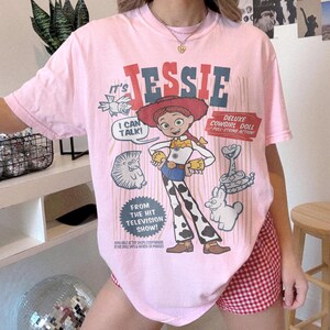 May include: Pink t-shirt featuring a graphic of Jessie, the cowgirl doll, from the television show. The design includes the text "JESSIE", "I CAN TALK!", and "DELUXE COWGIRL DOLL". The shirt has short sleeves.