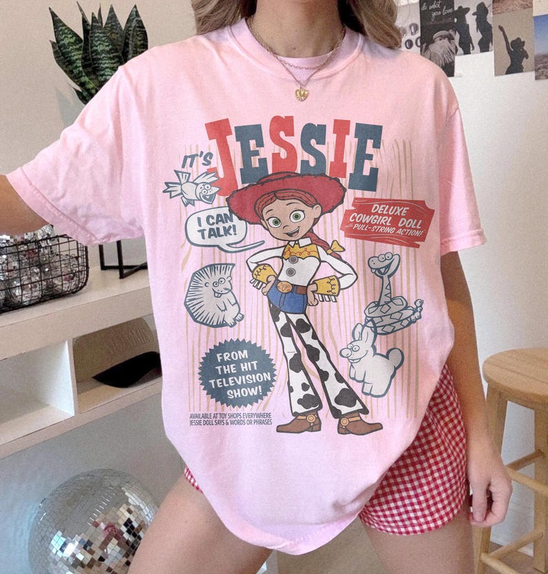 May include: Pink t-shirt featuring a graphic of Jessie, the cowgirl doll, from the television show. The design includes the text "JESSIE", "I CAN TALK!", and "DELUXE COWGIRL DOLL". The shirt has short sleeves.