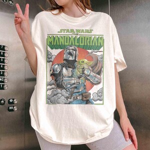 May include: Cream-coloured t-shirt with a graphic from "Star Wars: The Mandalorian." The design shows the Mandalorian character holding Grogu, set against a red sun and clouds. The text "Star Wars The Mandalorian" is displayed above the image.