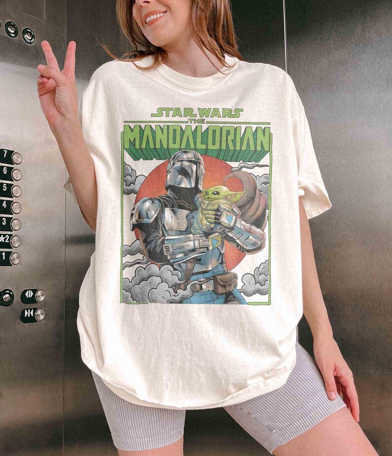 May include: Cream-coloured t-shirt with a graphic from "Star Wars: The Mandalorian." The design shows the Mandalorian character holding Grogu, set against a red sun and clouds. The text "Star Wars The Mandalorian" is displayed above the image.