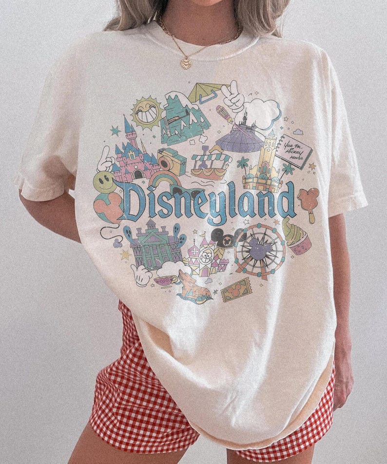 May include: Cream-colored t-shirt featuring a colorful Disneyland graphic. The design includes the word "Disneyland" in a blue font, surrounded by various illustrations of park attractions and characters. The shirt has short sleeves.