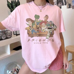 May include: A light pink short-sleeve t-shirt featuring a graphic print of various animated characters sitting on a couch. Below the characters, the text reads "F.R.I.E.N.D.S SINCE 1995" in a brown font. The shirt has a crew neckline and a relaxed fit.