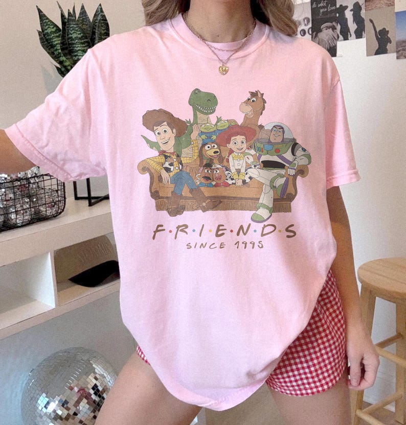 May include: A light pink short-sleeve t-shirt featuring a graphic print of various animated characters sitting on a couch. Below the characters, the text reads "F.R.I.E.N.D.S SINCE 1995" in a brown font. The shirt has a crew neckline and a relaxed fit.