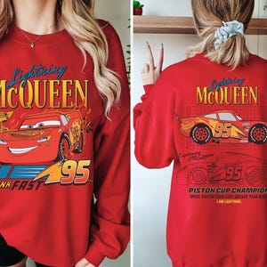 May include: Red sweatshirt featuring Lightning McQueen from the Cars movie. The front graphic shows McQueen with the number 95 and the text "THINK FAST". The back displays McQueen's image and the text "PISTON CUP CHAMPION".