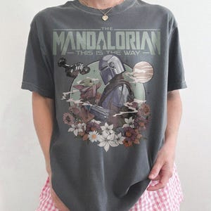 May include: Dark gray t-shirt featuring a graphic from "The Mandalorian" with the text "The Mandalorian - This is the Way." The design includes the Mandalorian, Grogu, and floral elements. The shirt is a relaxed fit.