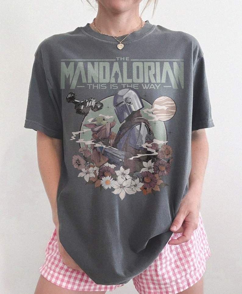 May include: Dark gray t-shirt featuring a graphic from "The Mandalorian" with the text "The Mandalorian - This is the Way." The design includes the Mandalorian, Grogu, and floral elements. The shirt is a relaxed fit.