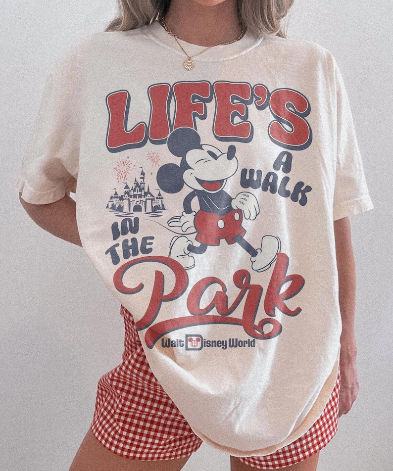 May include: Cream-colored t-shirt featuring a graphic of Mickey Mouse walking with the text "LIFE'S A WALK IN THE PARK" in red and blue. The shirt also includes the text "Walt Disney World".