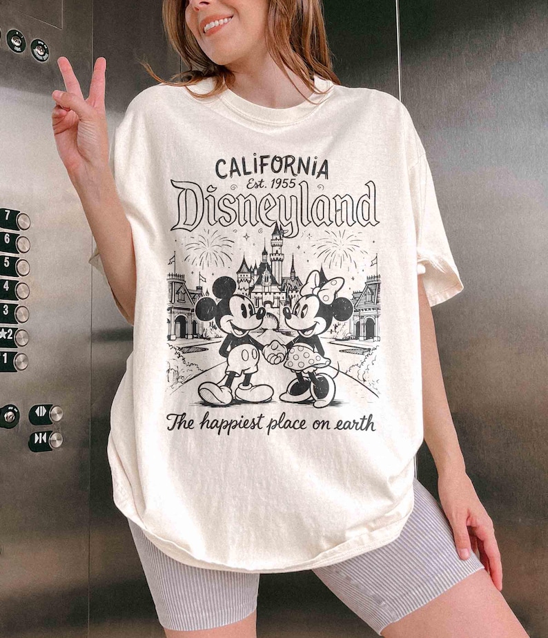 May include: Cream-coloured t-shirt with a black and white graphic of Mickey and Minnie Mouse in front of Disneyland. The text "CALIFORNIA Disneyland Est. 1955" and "The happiest place on earth" are printed on the shirt.