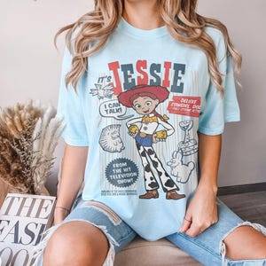 May include: Light blue t-shirt featuring a graphic of the character Jessie from the Toy Story franchise. The design includes the text "Jessie", "I Can Talk!", and "Deluxe Cowgirl Doll". The shirt is a casual wear item.