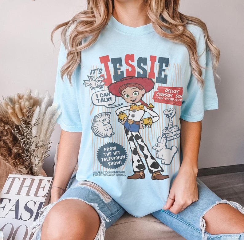 May include: Light blue t-shirt featuring a graphic of the character Jessie from the Toy Story franchise. The design includes the text "Jessie", "I Can Talk!", and "Deluxe Cowgirl Doll". The shirt is a casual wear item.