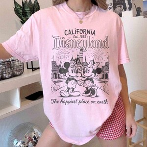 May include: Pale pink t-shirt featuring a Disneyland graphic. The design includes Mickey and Minnie Mouse, a castle, fireworks, and the text "California Disneyland Est. 1955" and "The happiest place on earth."