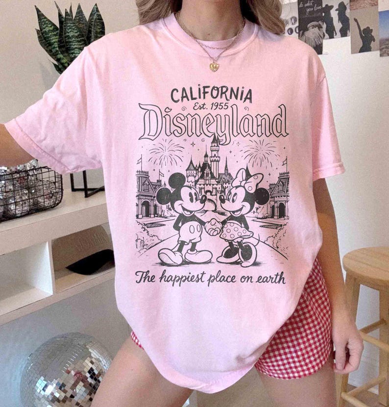 May include: Pale pink t-shirt featuring a Disneyland graphic. The design includes Mickey and Minnie Mouse, a castle, fireworks, and the text "California Disneyland Est. 1955" and "The happiest place on earth."