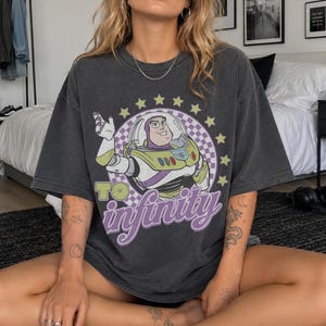 May include: A charcoal gray short-sleeve t-shirt featuring a graphic of Buzz Lightyear inside a purple and white checkered circle surrounded by yellow stars. The text "TO infinity" is printed in large, bold, light green and purple lettering.