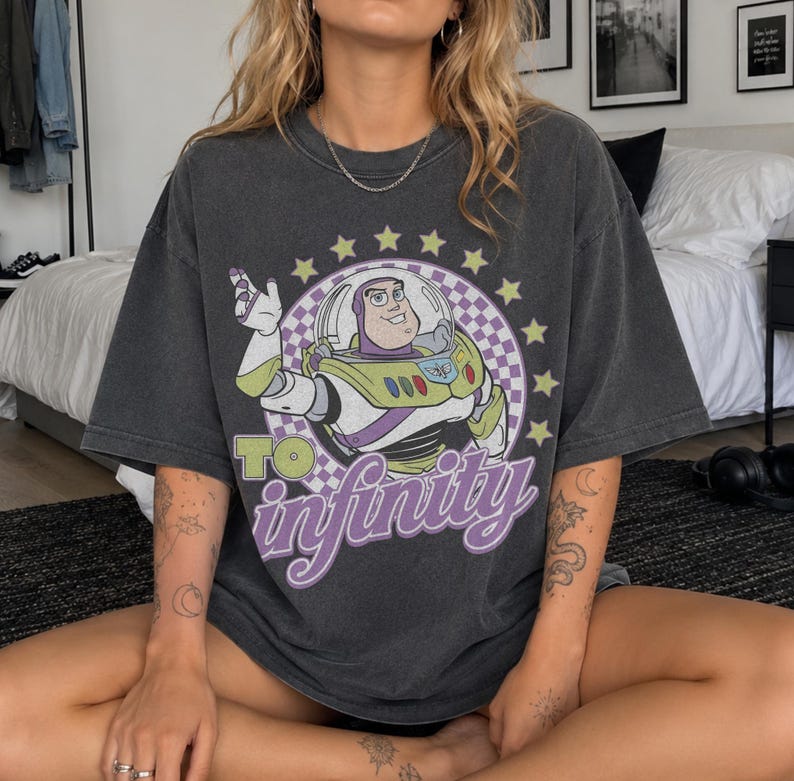 May include: A charcoal gray short-sleeve t-shirt featuring a graphic of Buzz Lightyear inside a purple and white checkered circle surrounded by yellow stars. The text "TO infinity" is printed in large, bold, light green and purple lettering.
