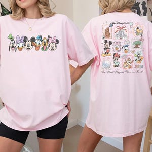 May include: A light pink t-shirt featuring Disney characters' faces across the front. The back of the shirt displays a collage of Disney World-themed images and text, including "Walt Disney World" and "The Most Magical Place on Earth."