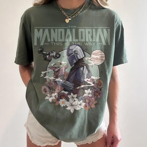 May include: Olive green t-shirt featuring a graphic of The Mandalorian and Grogu surrounded by flowers, with the text "THE MANDALORIAN - THIS IS THE WAY." The design includes a helmeted figure and a small, green alien. The shirt is a comfortable fit.