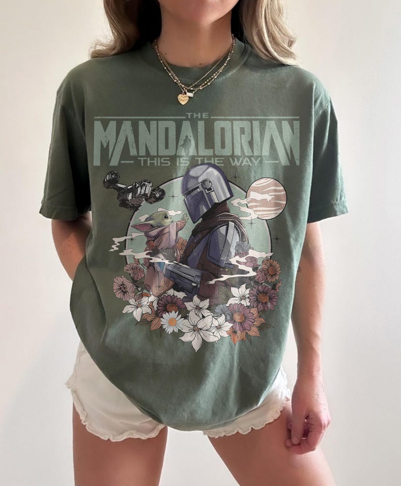 May include: Olive green t-shirt featuring a graphic of The Mandalorian and Grogu surrounded by flowers, with the text "THE MANDALORIAN - THIS IS THE WAY." The design includes a helmeted figure and a small, green alien. The shirt is a comfortable fit.