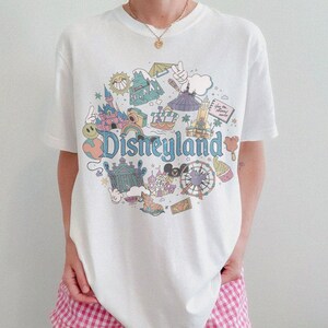May include: White t-shirt with a colorful Disneyland graphic. The design features iconic park elements like a castle, a Ferris wheel, and the Disneyland logo in a retro style. The shirt is a classic crew neck.