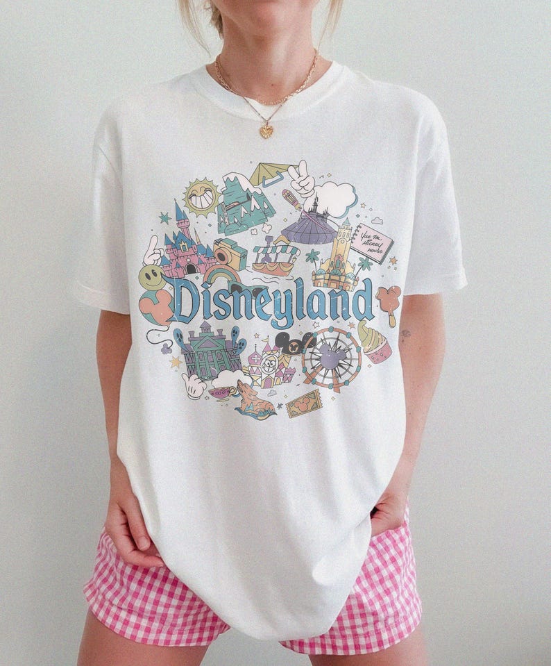 May include: White t-shirt with a colorful Disneyland graphic. The design features iconic park elements like a castle, a Ferris wheel, and the Disneyland logo in a retro style. The shirt is a classic crew neck.