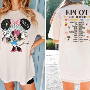 May include: Cream-colored t-shirt featuring a Minnie Mouse graphic with a pink bow, blue dress, and a wand. The text "2026" is above the character. The back of the shirt displays "EPCOT WORLD TOUR" and various city names.