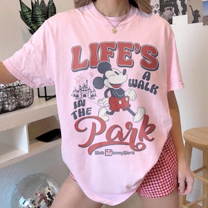 May include: A light pink t-shirt featuring Mickey Mouse walking in front of a castle with the text "LIFE'S A WALK IN THE PARK" and "Walt Disney World" in red and white. The shirt is a crew neck.