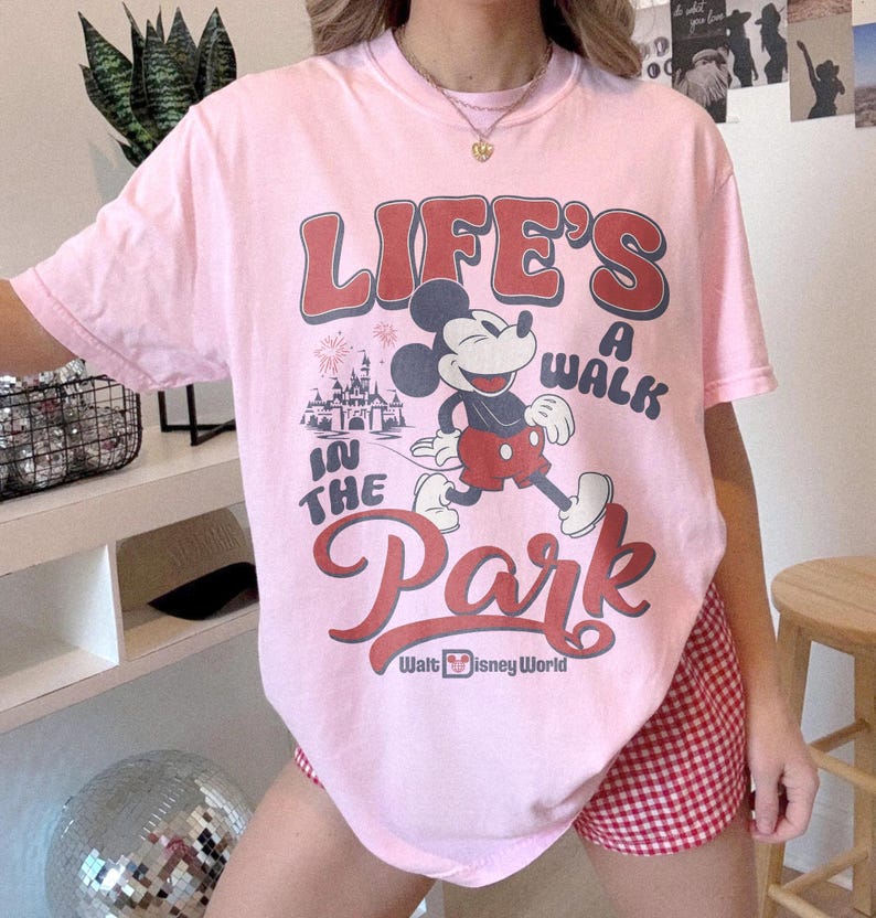May include: A light pink t-shirt featuring Mickey Mouse walking in front of a castle with the text "LIFE'S A WALK IN THE PARK" and "Walt Disney World" in red and white. The shirt is a crew neck.