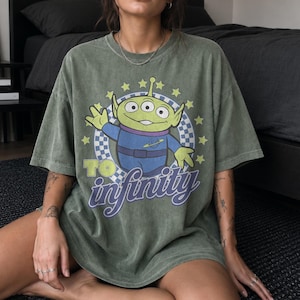 May include: An olive green short-sleeve t-shirt featuring a graphic of a three-eyed green alien character in a blue space suit. The design includes a checkered circle, yellow stars, and the text TO infinity in purple and yellow lettering.