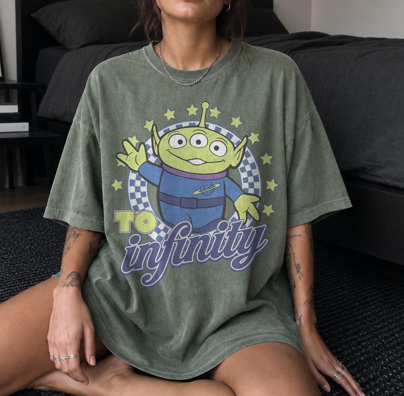 May include: An olive green short-sleeve t-shirt featuring a graphic of a three-eyed green alien character in a blue space suit. The design includes a checkered circle, yellow stars, and the text TO infinity in purple and yellow lettering.