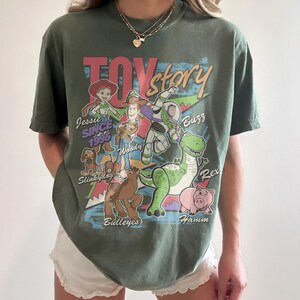 May include: An olive green short-sleeve t-shirt featuring a colorful Toy Story graphic print. The design includes characters Jessie, Woody, Buzz Lightyear, Slinky Dog, Bullseye, Rex, and Hamm with their names and the text Toy Story Since 1995.