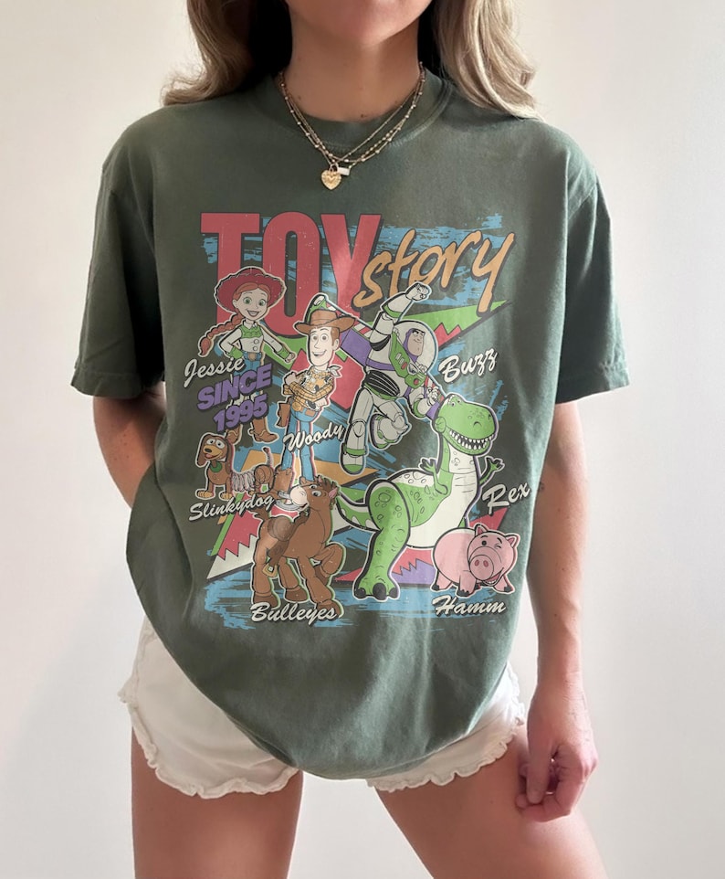 May include: An olive green short-sleeve t-shirt featuring a colorful Toy Story graphic print. The design includes characters Jessie, Woody, Buzz Lightyear, Slinky Dog, Bullseye, Rex, and Hamm with their names and the text Toy Story Since 1995.