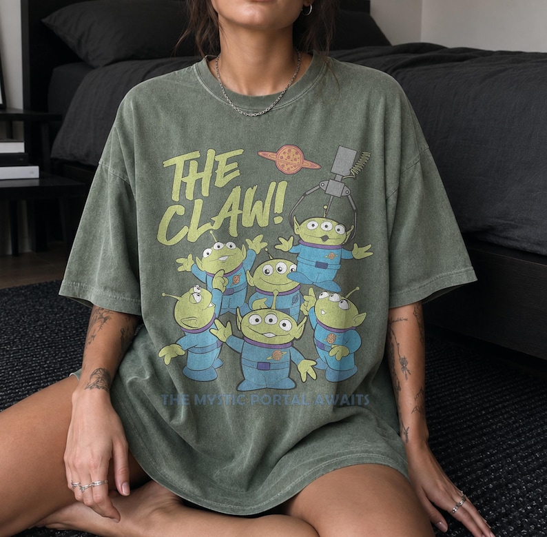 Puede incluir: Una camiseta de manga corta color verde oliva con un estampado gr&aacute;fico de cinco alien&iacute;genas verdes de tres ojos con trajes espaciales azules. Arriba, el texto dice THE CLAW! con un icono de planeta. Debajo, el texto dice THE MYSTIC PORTAL AWAITS.